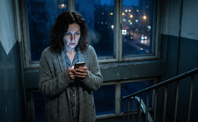 A woman reads a midnight message from debt collectors on social media in a dark apartment stairwell.