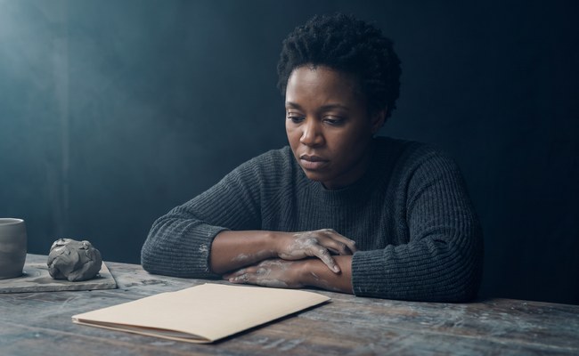 A potter stops work at her bench and looks down at a closed folder beside the clay, her expression falling.