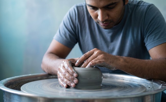 A potter centers a ball of grey clay on a spinning wheel with water running down his wrists.