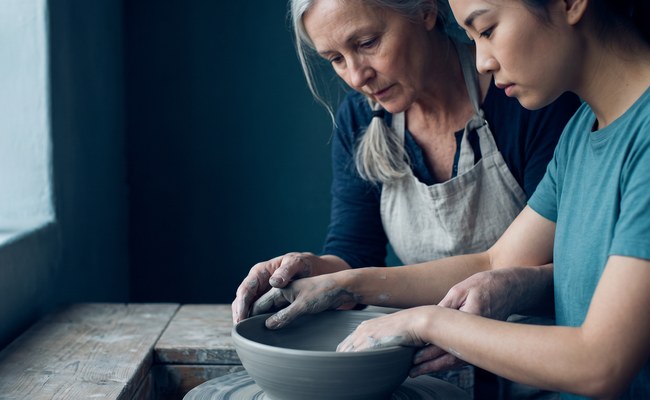 An experienced potter steadies the hands of a younger one as she shapes the rim of a clay bowl.