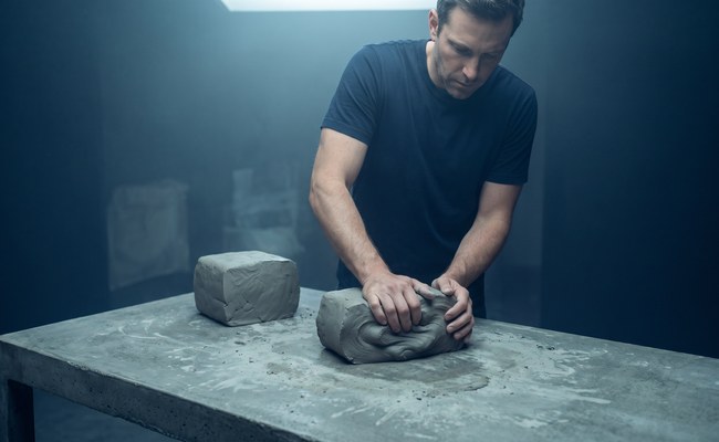 A potter wedges a block of grey clay on a concrete table beside an identical block already prepared.