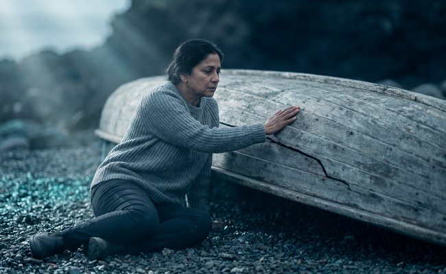 A woman kneels beside an overturned wooden dinghy with her hand resting on a crack in the hull, no longer working.
