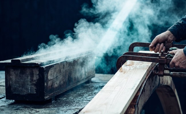 A timber steam box vents white vapor as a softened plank is bent around a form and held with iron clamps.
