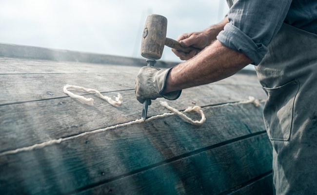 A caulking iron and wooden mallet drive cotton into a single seam between two planks on a boat hull.
