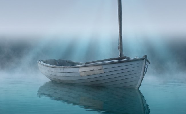 A repaired wooden dinghy lies quietly afloat at a mooring in cool pre-dawn light with mist rising off the water.
