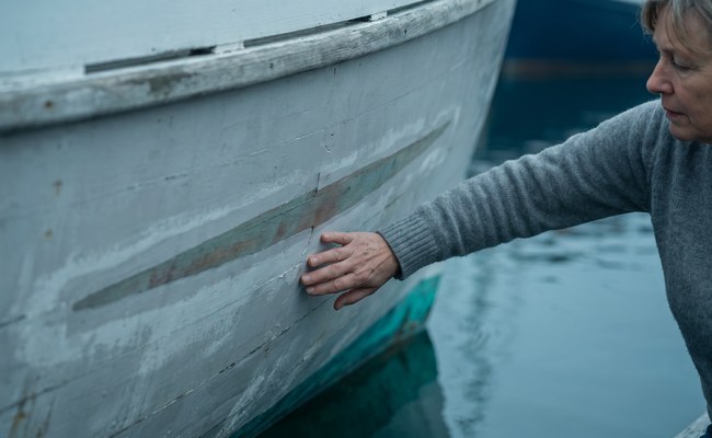 A woman rests an open hand on a boat hull where an old repair is still faintly visible beneath fresh primer.
