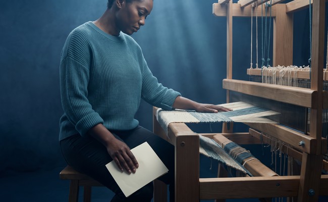 A weaver stops work at her loom holding a letter face-down after her credit report dispute was denied.
