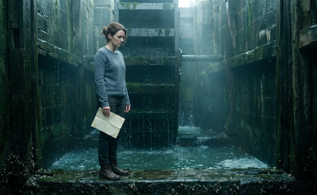 A tide mill keeper stands in a stone wheel pit holding a letter she has just read, her shoulders dropped.
