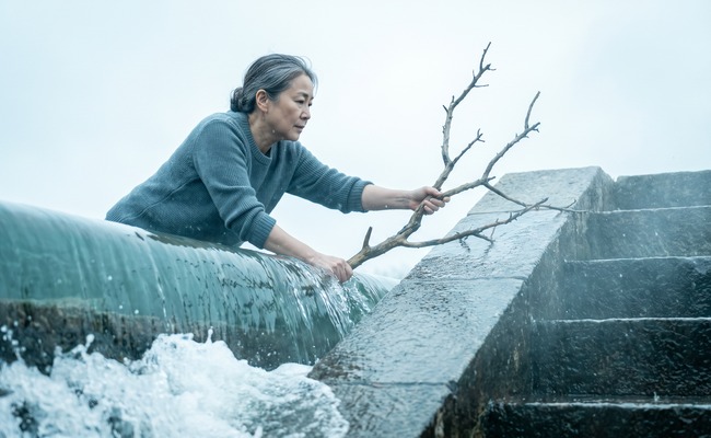 A woman leans over a stone weir to lift a caught branch out of the water sheeting across the crest.

