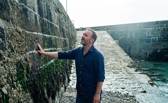 A man rests his hand against a slipway wall banded with weed and barnacle growth marking the levels the water repeatedly reaches.
