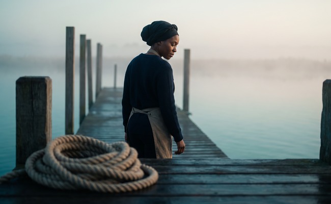 A woman walks a timber pier over flat water at dawn, glancing down at the surface as she passes a coil of mooring rope.

