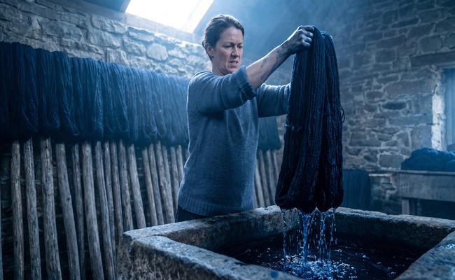 A woman lifts a heavy sodden skein of indigo-dyed wool from a vat with bare hands stained blue to the wrist.