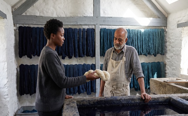 A woman holds out a single undyed skein toward an older man across a dye trough, and he looks at it with consideration.