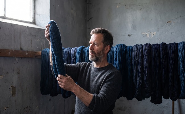 A man holds one finished skein of evenly dyed indigo wool up to the light while dozens more hang on the rod behind him.