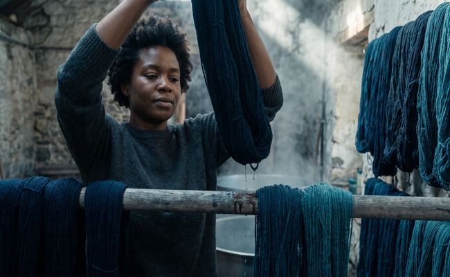 A woman hangs a freshly dyed navy skein onto a drying rod, water still running from the wool, steam catching the light.