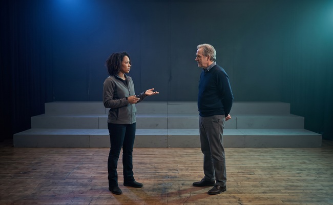 Two adults calmly discuss what a credit-card late-fee waiver can and cannot change on an empty rehearsal stage.