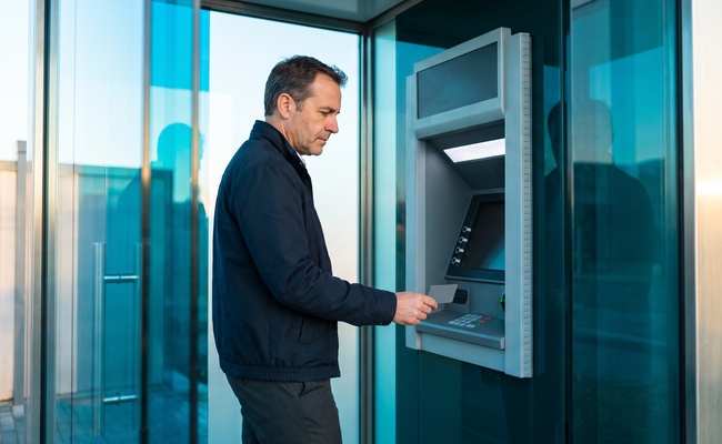 A person checks a plain card and dark ATM display inside a bright glass vestibule before taking cash.