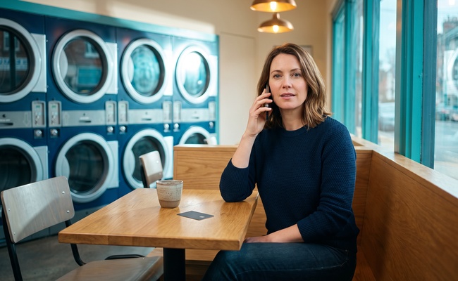 A person makes a careful budget decision by phone in a bright laundromat seating nook after an account closure.