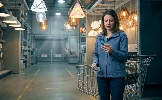 Rachel pauses beside an empty shopping cart after learning her credit card limit was lowered, checking a plain card and phone in a bright lighting aisle.