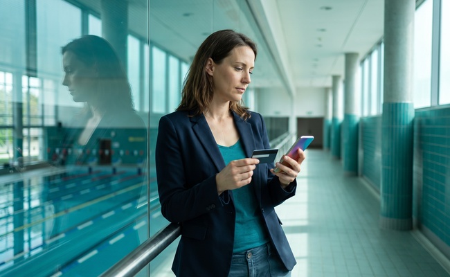 A person adult person compares a plain card and abstract phone beside an empty aquatic-center corridor while identifying a credit-card billing error.