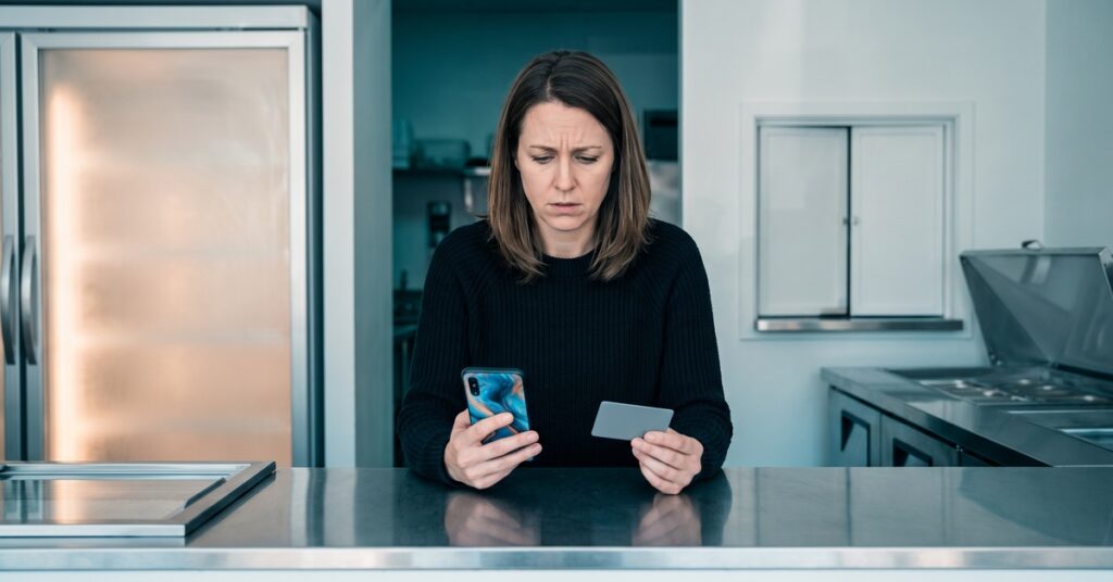 A person adult person checks an abstract phone behind a blank stainless ice-cream-shop service counter beside a closed freezer door after a credit card chargeback vs refund decision remains unresolved.