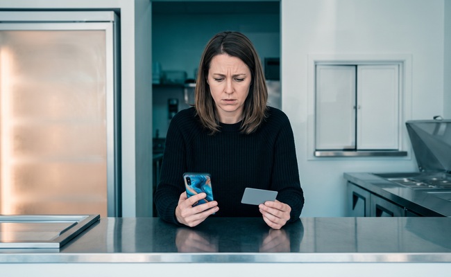 A person studies an abstract phone behind a blank stainless ice-cream-shop service counter while a credit card chargeback vs refund choice remains unresolved.
