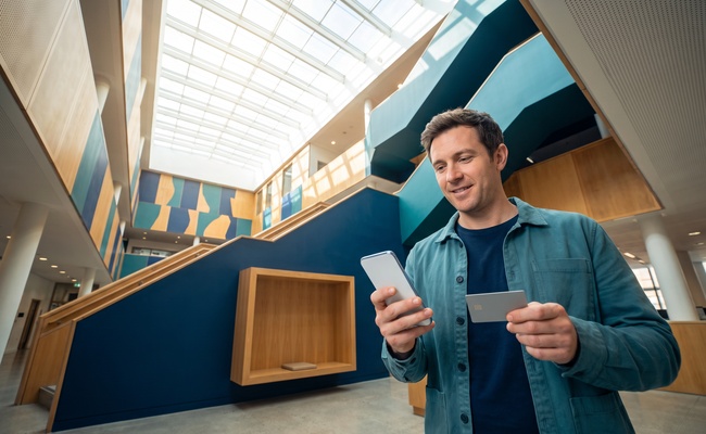 A person makes a careful payment through a blank phone in a bright library atrium, leaving a cushion as he prepares to pay before the statement closes.