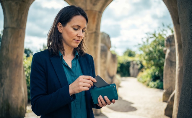A person checks a blank phone and secures a plain card in a sculpture-garden pavilion after reviewing the result of choosing to pay before the statement closes.