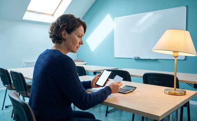 A person checks a blank phone and plain card in an adult-education classroom while verifying a credit card overpayment in the account history.