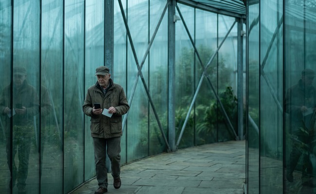 A person older adult person investigates a pending credit card payment on a blank phone while walking through a glass conservatory.