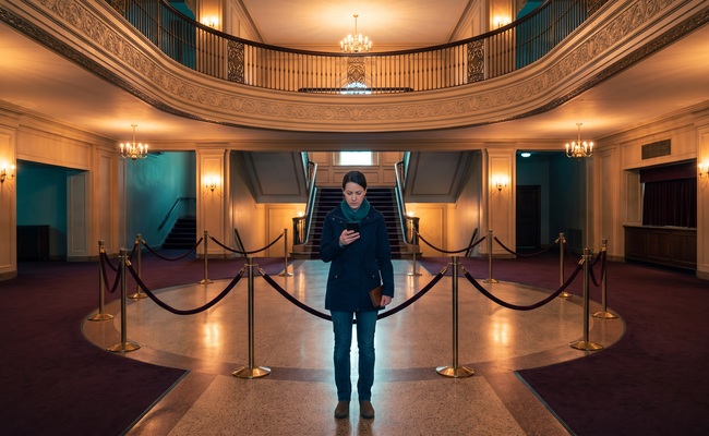 A person adult person checks personal loan default credit-report information in a civic theater lobby with a blank phone and closed wallet.