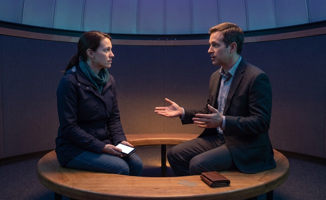 A person and an adult adviser discuss a personal loan default in an observatory vestibule using blank phones and a closed wallet.
