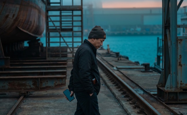 A person adult person considers co signer loan risks while walking beside an unbranded harbor dry dock with a blank phone and closed wallet.