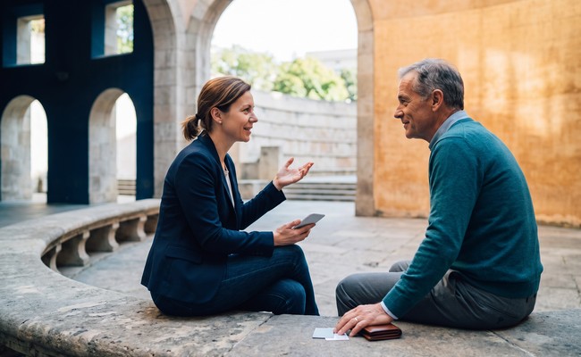 Two adults discuss payoff quotes, reserves, and whether to pay off personal loan early in a sunlit civic courtyard.