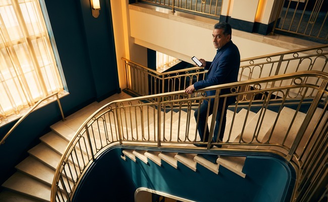 an adult Latino person in his early 60s considers how to refinance a personal loan in a art-deco hotel staircase with sweeping rails and a landing visible above.