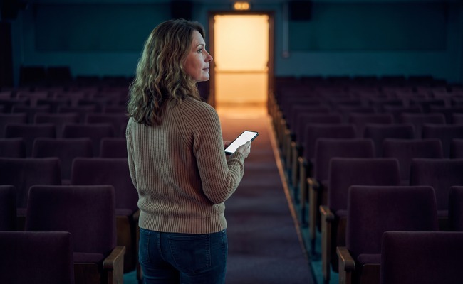 an adult person in her early 50s considers how to refinance a personal loan in a quiet independent cinema aisle with rows of unmarked seats and a bright exit glow.