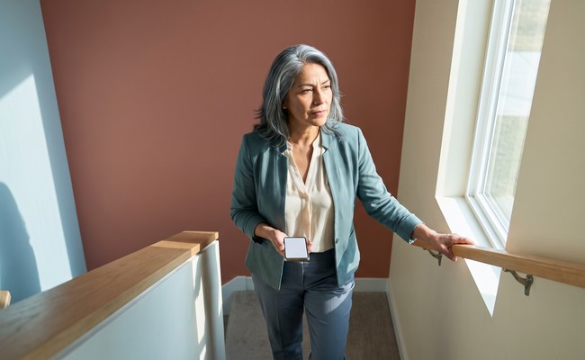 an adult Latino person in her late 50s responds to car loan repossession warning signs in a sunlit residential stair landing with split-level geometry and an open passage.