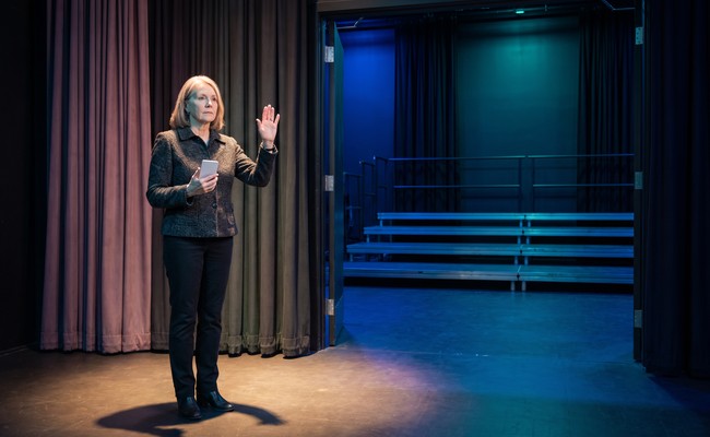an adult person in her late 60s responds to what to do after car repossession in a unbranded person-box theater stage with empty risers, curtain wings, and deep axial space.