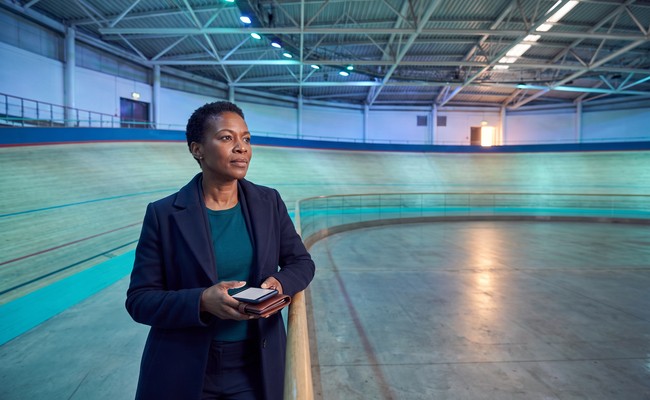 an adult person in her early 50s responds to what to do after car repossession in a indoor velodrome with sweeping track curves, open infield, and high roof trusses.