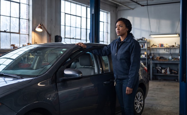 A person the emotional hook: a person pauses beside a car repair bay, weighing a major repair and a careful cash plan.