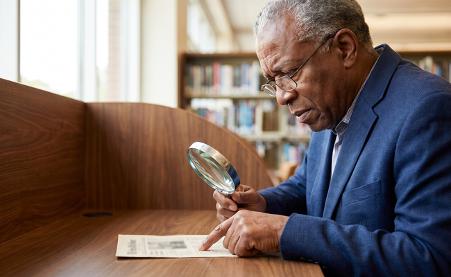 Older man using a magnifier to inspect an unreadable statement at a library study carrel.