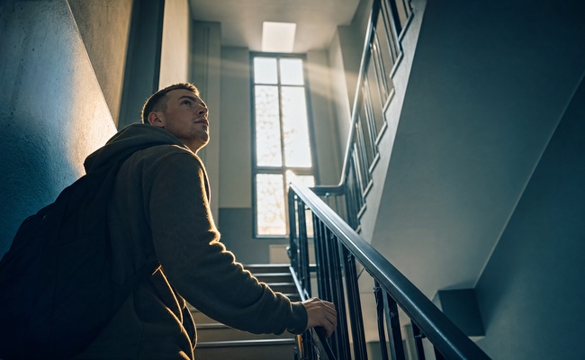 A young man pauses on a bright stairwell landing and looks upward while deciding which path to take.