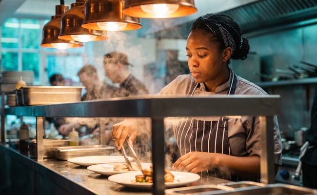 Young cook plating food with focused competence at a busy commercial kitchen pass line.