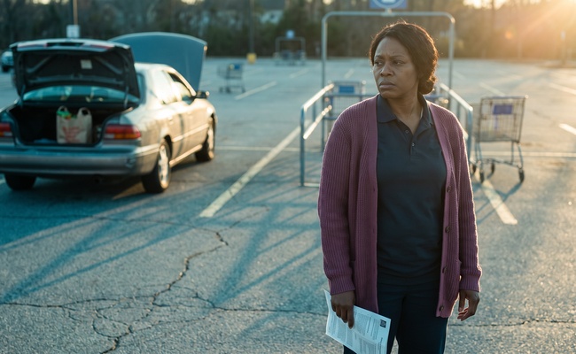 A woman stands beside her broken-down car in a grocery store parking lot holding a repair estimate at her side.