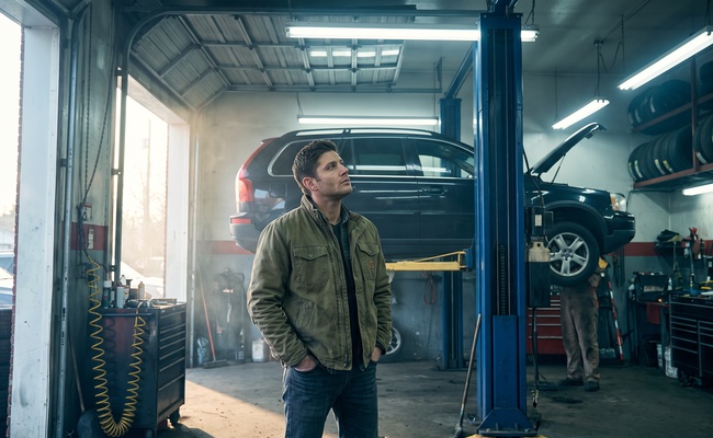A man stands in an auto repair bay looking up at his car on a lift while waiting for the cost of the repair.