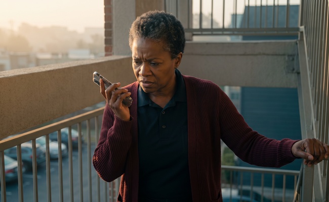A woman in a maroon cardigan stands on an apartment stairwell at dawn holding her phone away from her ear with a worried expression.