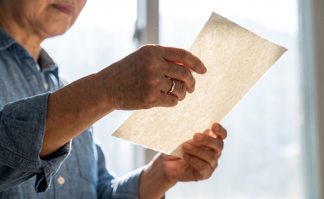 Close-up of weathered hands holding a single sheet of paper up toward bright window light.