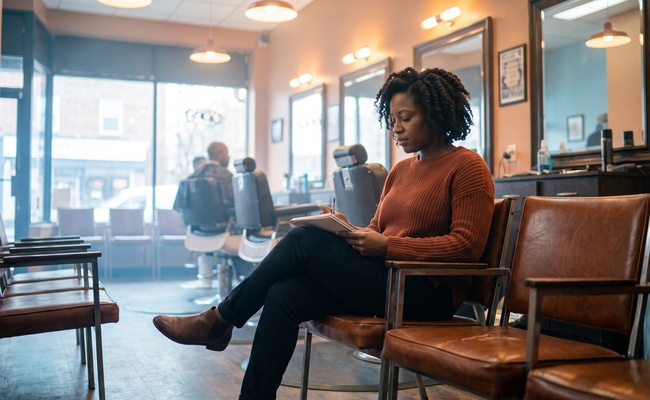 A woman sits in a barbershop waiting chair reading a page on her knee with a pen ready in her hand.