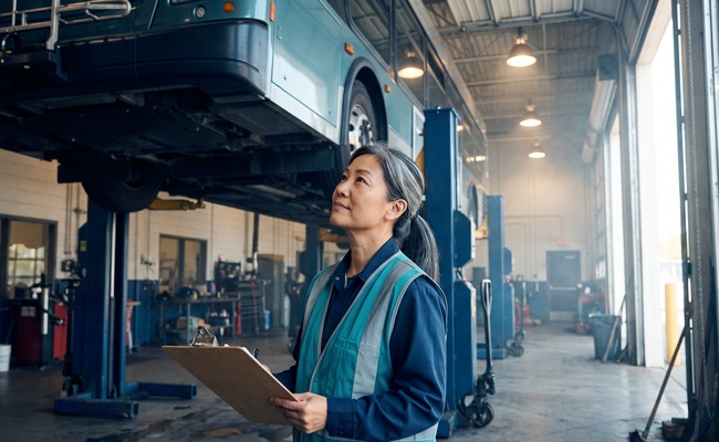 Transit inspector holding a clipboard while checking a record against the vehicle above her.