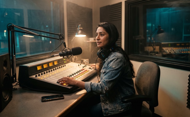 Woman at a radio station mixing console at night with her phone lying face down beside her.